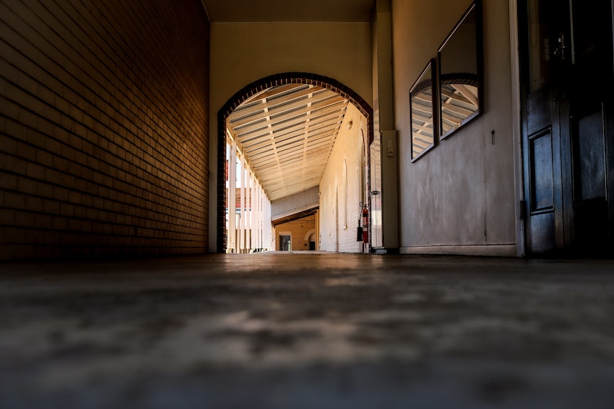 An indoor corridor with a brick wall on one side and a wooden door on the other looking toward an outside walkway