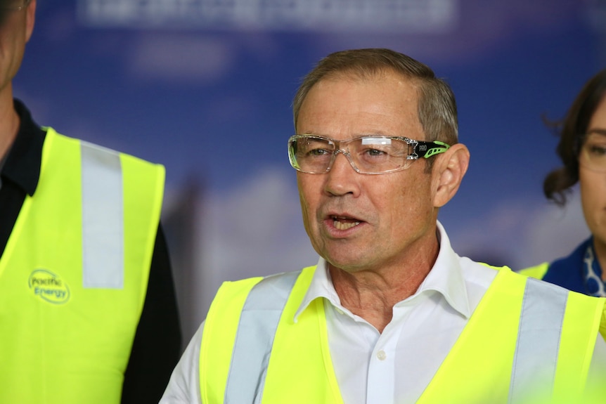 A man wearing a high-vis vest and safety glasses speaks at a press conference.