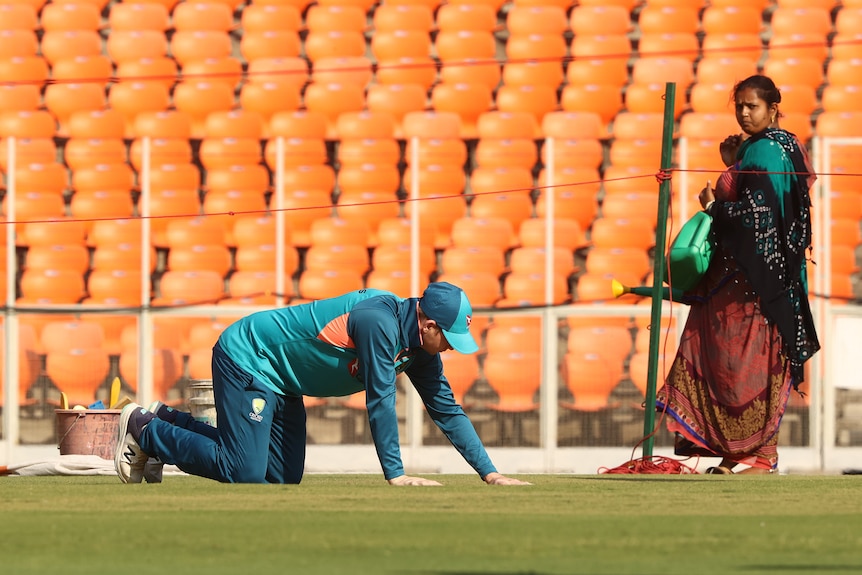 Steve Smith on all fours rubs his hand across the grass at Narendra Modi Stadium. A woman with a watering can watches him.