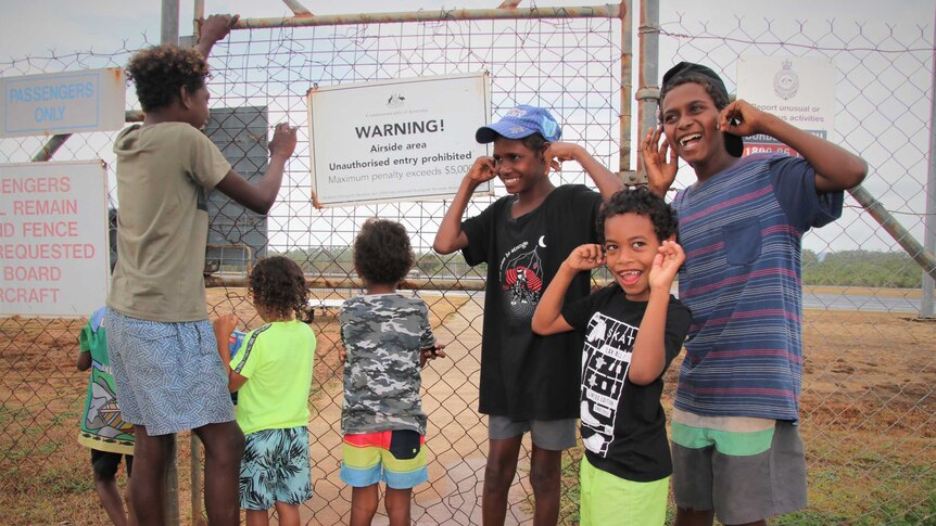 Aboriginal children, some blocking their ears, standing in front of a fence in an airside area
