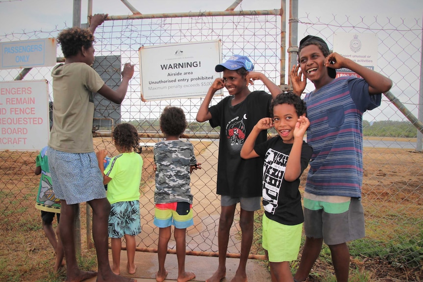 Aboriginal children, some blocking their ears, standing in front of a fence in an airside area