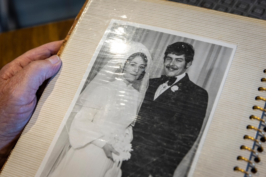 A black and white wedding photo in an album, the bride and groom are smiling dressed in 1970s wedding attire.