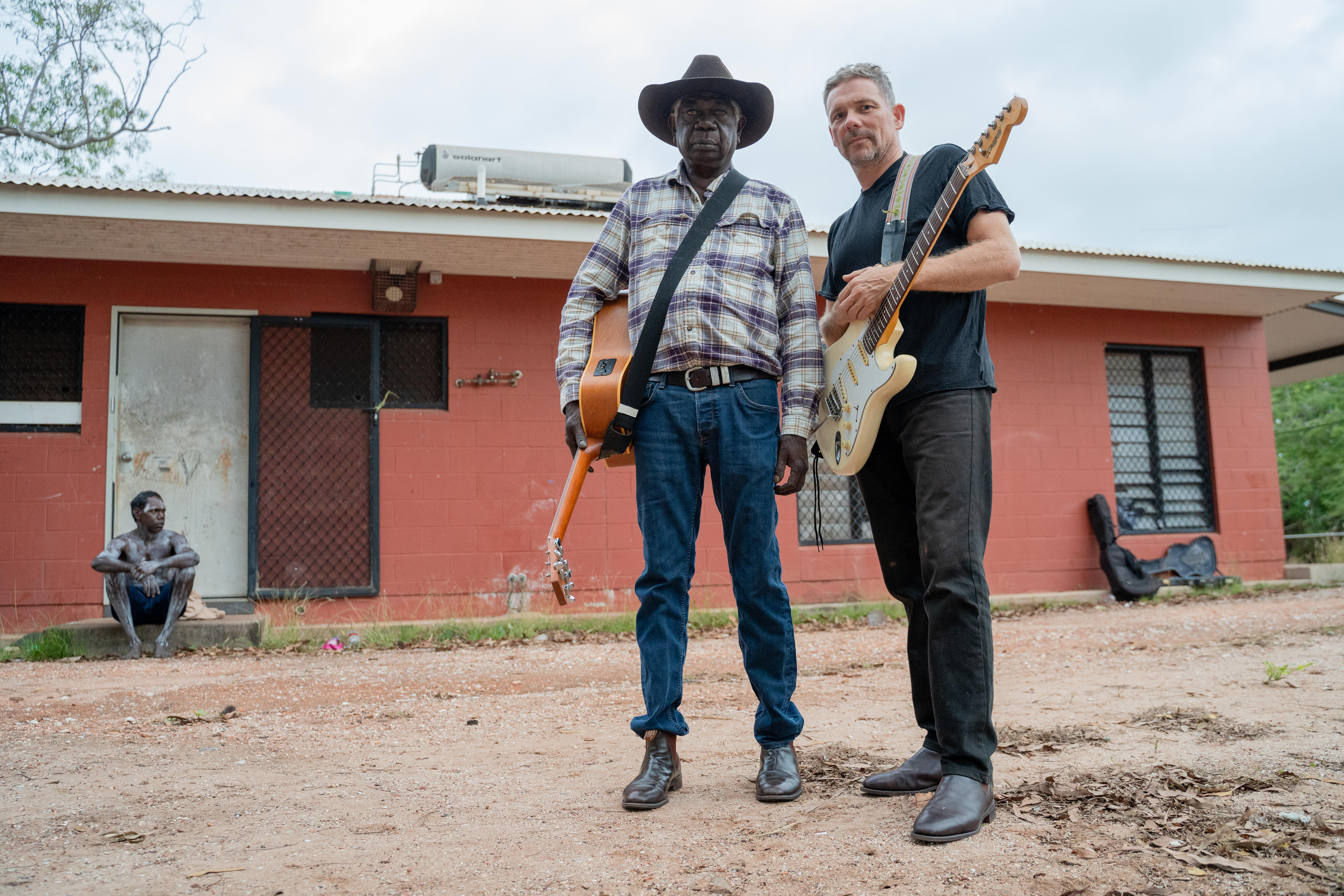 Two men are holding guitars and standing outside a red house. The house has a man sitting in front of a closed white door. 