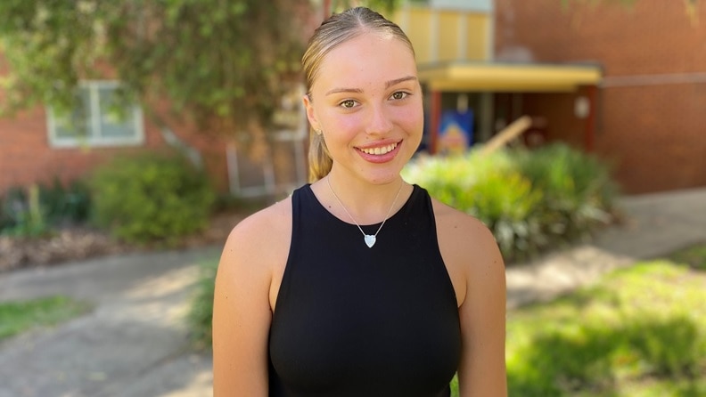 A young girl with blonde ponytail, black sleeveless top stands in front of red building, plants, smiles at the camera.