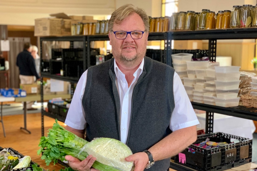 a middle-aged man holds green vegetables with shelves of dry goods behind him