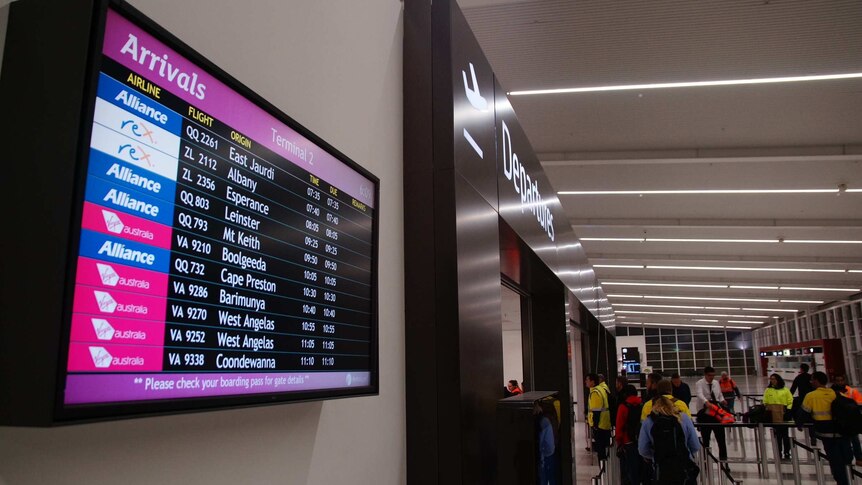 The arrivals board at Perth Airport showing the flights arriving from a range of mining towns, as workers queue at the counter.