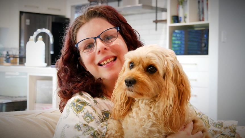 A woman with curly hair and glasses smiling and cuddling a dog.