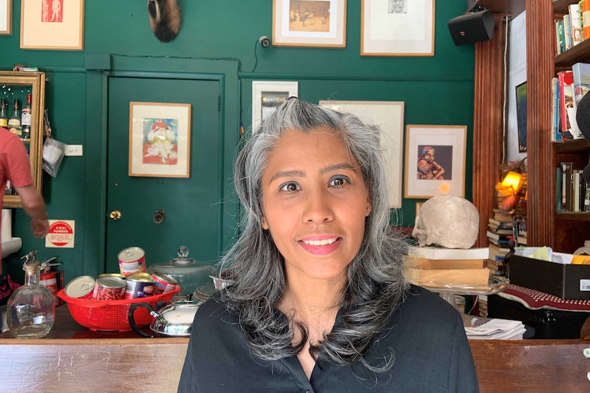 A woman sits in a room with a green wall covered in framed artworks and a bookshelf