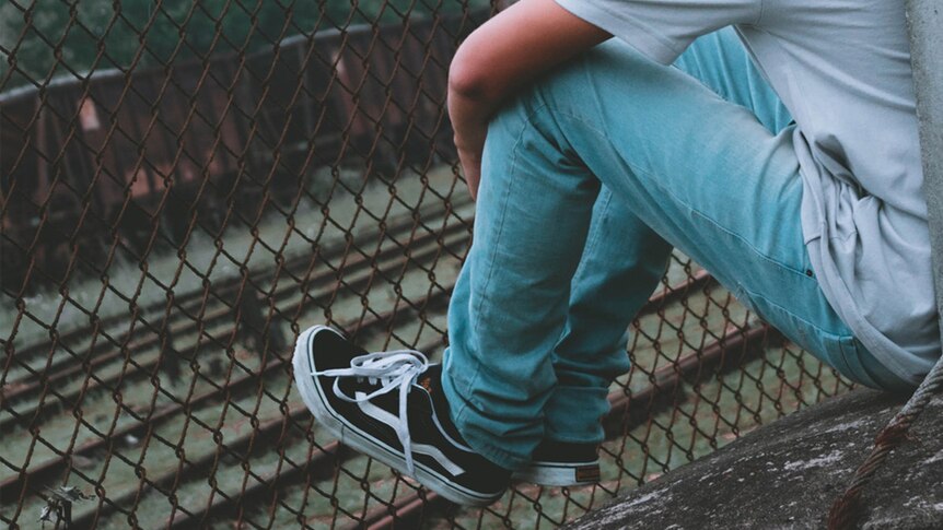 A young man in blue jeans rests his sneakers on a rusty fence