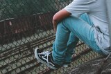 A young man in blue jeans rests his sneakers on a rusty fence