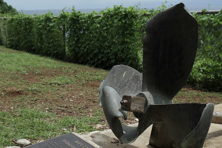 A monument overlooking a beach features a plaque and broken propeller