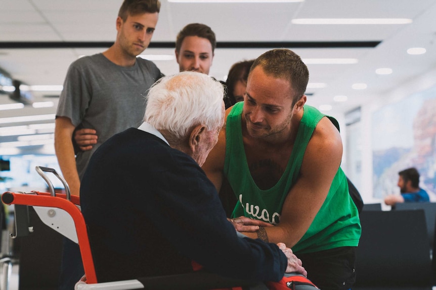 A young man in a green singlet hugs an old man in a wheelchair as other family look on.