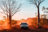 A four wheel drive travels down a dirt road as the sun sets in the background.