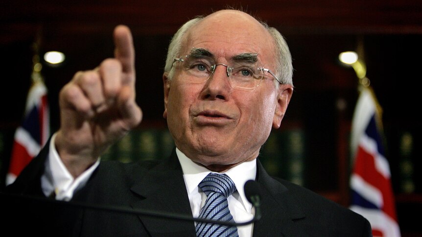 A close-up photo of John Howard pointing to an audience, with Australian flags in the background