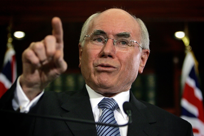 A close-up photo of John Howard pointing to an audience, with Australian flags in the background