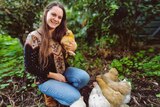 A woman squats down in her garden, with four chickens at her feet and one in her arms.