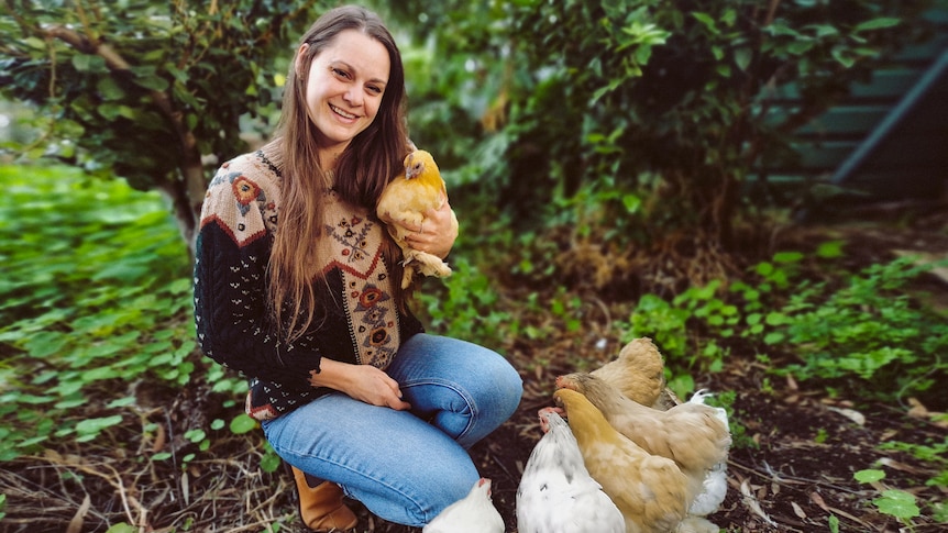 A woman squats down in her garden, with four chickens at her feet and one in her arms.