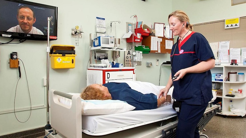 A hospital treatment room with a  patient on a bed, nurse by her side, and doctor appearing on a large TV screen on the wall