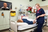 A hospital treatment room with a  patient on a bed, nurse by her side, and doctor appearing on a large TV screen on the wall