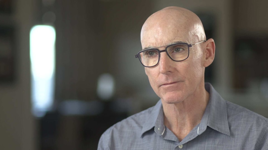 A bald man with thick black rimmed glasses wearing a blue open collared shirt with the background out of focus.