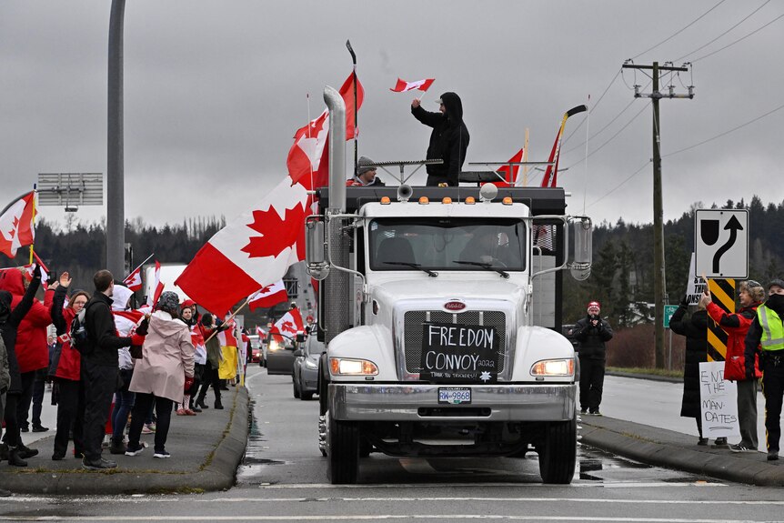 A large white truck adorned with Canadian flags drives along a road. People in the footpath wave flags as it passes