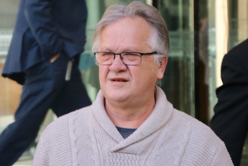 A head and shoulders shot of an older man wearing spectacles and a beige jumper outside court.