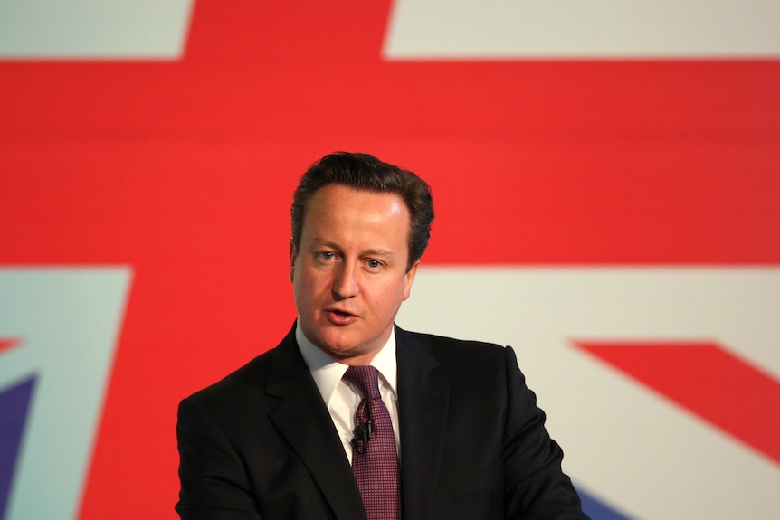 A clean-shaven man wearing a suit and dark red tie in front of a giant Union Jack.