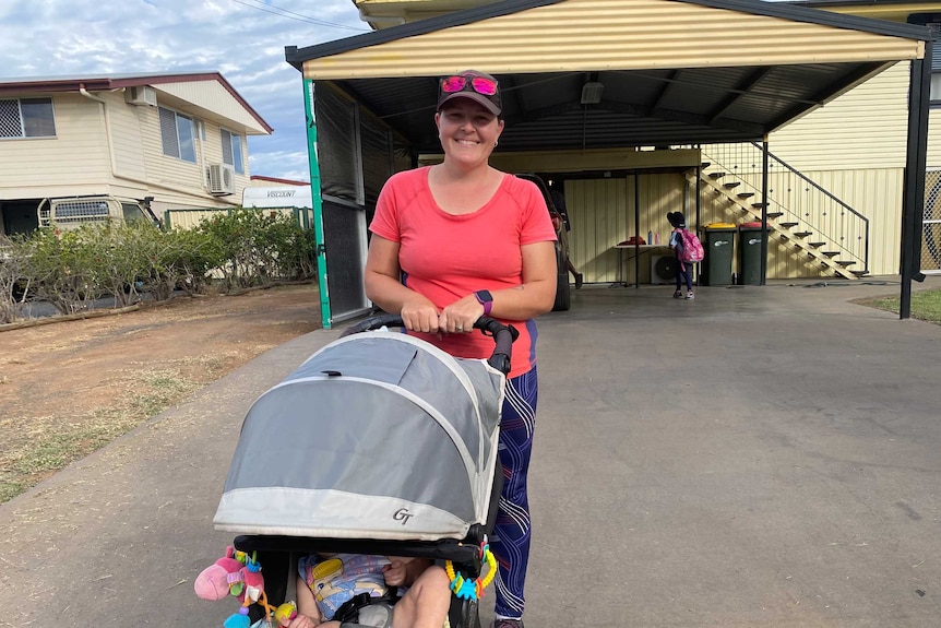 A woman in activewear and a cap pushing a pram with a baby inside while children play on the driveway of a house behind her