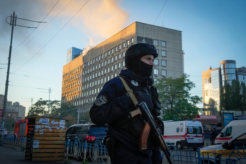 A man in a dark blue police uniform carrying a rifle and wearing a balaclava stands in front of a smoking building.