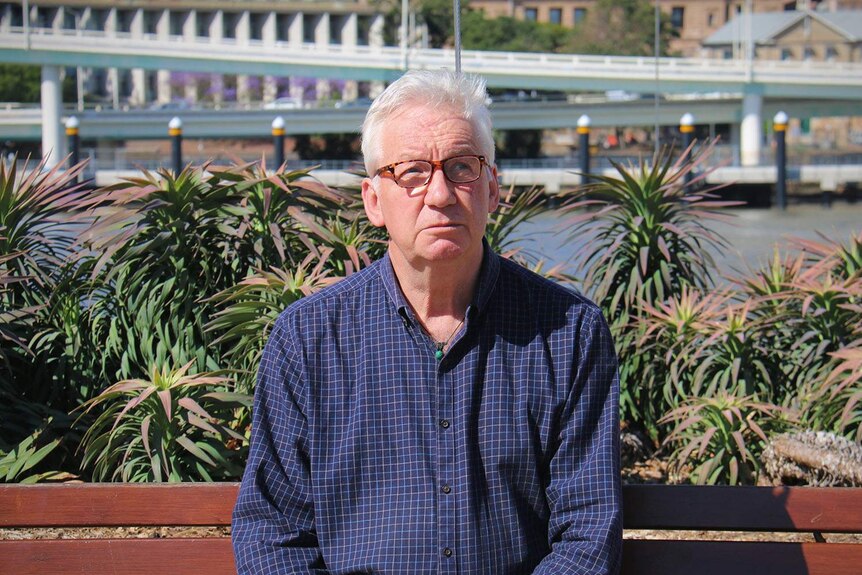 Professor John Wanna sits outside at South Bank with Brisbane river and city behind him.