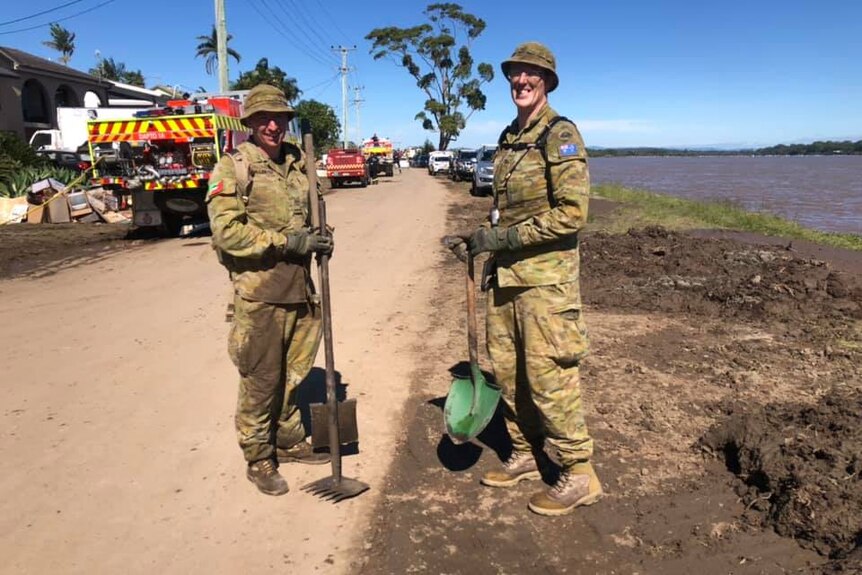 Two young men in Defence uniforms with shovels covered in mud on a dirt road.