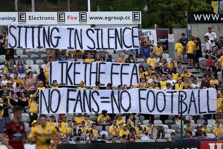 Soccer fans wearing yellow jerseys hold up a protest sign in the stands during a game