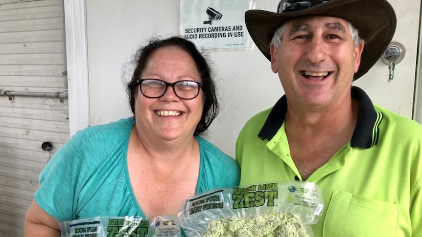 A couple smile at the camera holding up bags of lime zest with a closed door and camera security sign up behind them.