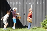 Three boys wearing bunny rabbit ear hop in front of a shed with 'Happy Easter' written on it.