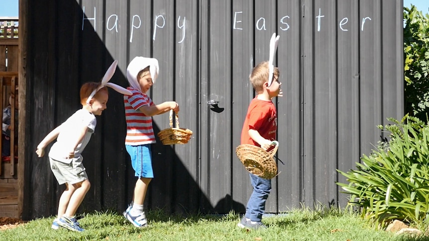 Three boys wearing bunny rabbit ear hop in front of a shed with 'Happy Easter' written on it.