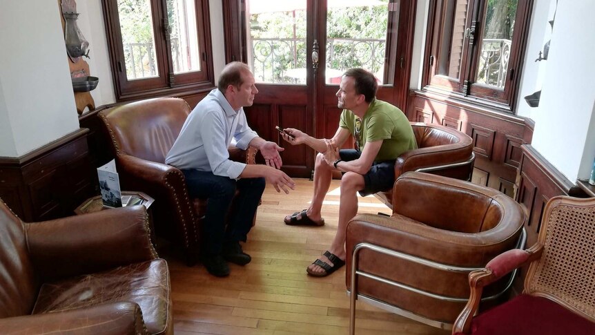 Christian Prudhomme leans in to talk to Rob Arnold, right, who records on his phone as they sit down on leather chairs.