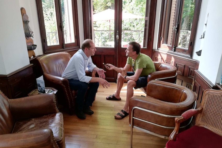 Christian Prudhomme leans in to talk to Rob Arnold, right, who records on his phone as they sit down on leather chairs.