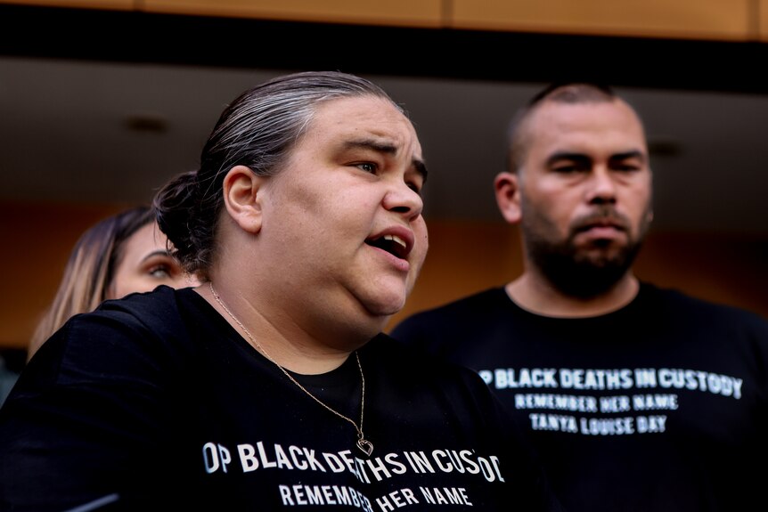 Woman with black and grey hair, wearing a t-shirt that reads No Black Deaths in Custody, with a man standing behind.