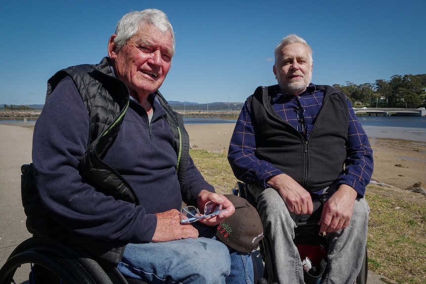 Two older men in wheel chairs sit side-by-side in front of grass and a lake, smiling at the camera