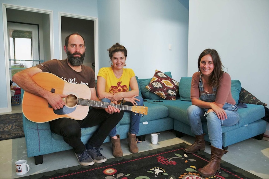 Three people, one of them with a guitar, sit on a couch.