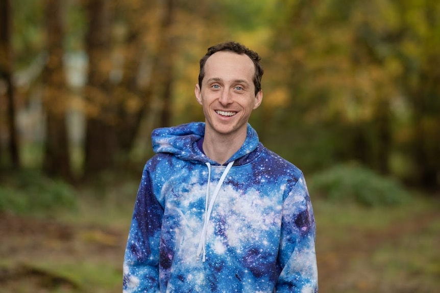 A young man smiles at the camera with a forest landscape in the background