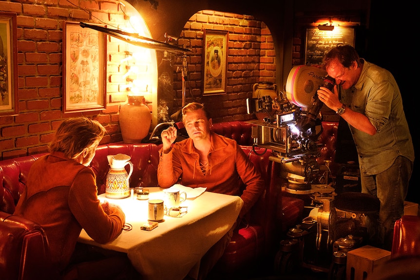 Two men sit in a booth, one smoking, face each other as another man to their right looks through cinema camera and films them.