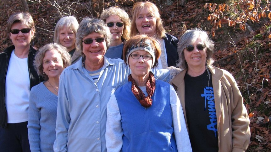 A group of eight women casually dressed stand close together, smiling and looking towards camera, with scrub in the background.