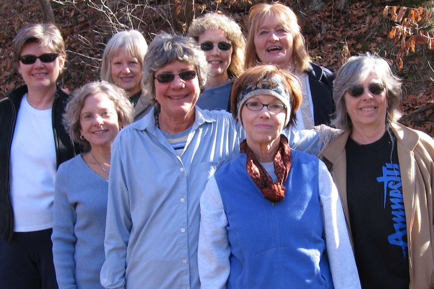 A group of eight women casually dressed stand close together, smiling and looking towards camera, with scrub in the background.