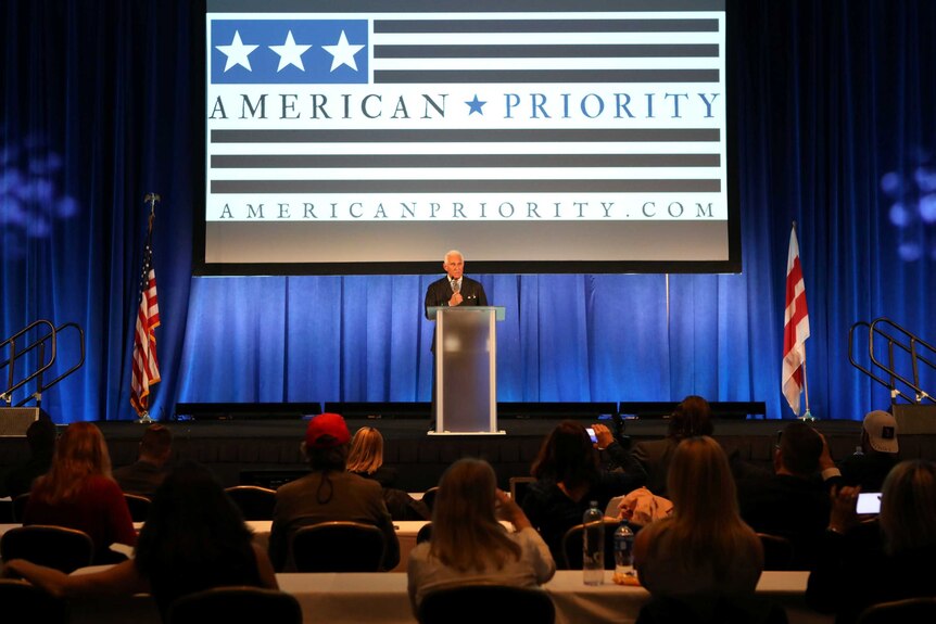 Roger Stone speaks in front of a podium with a large screen behind him