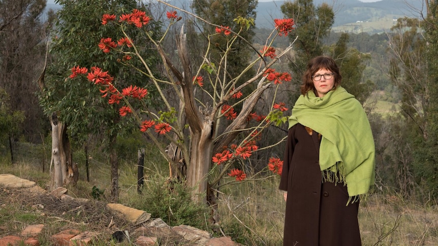 A woman stands in the bush, with a tree with bright red flowers behind her. 