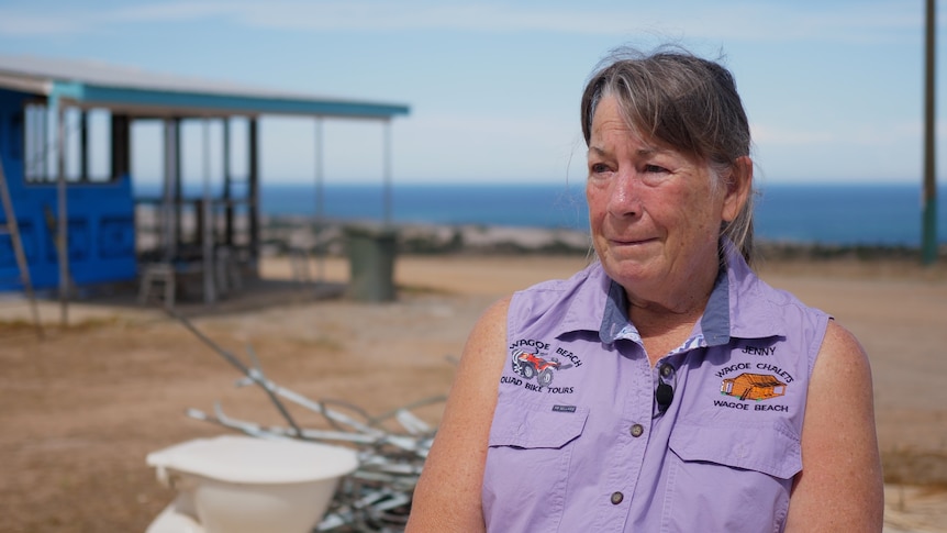 A woman in a purple blouse with tears in her eyes. Behind her is a toilet, twisted metal and the shell of a building.