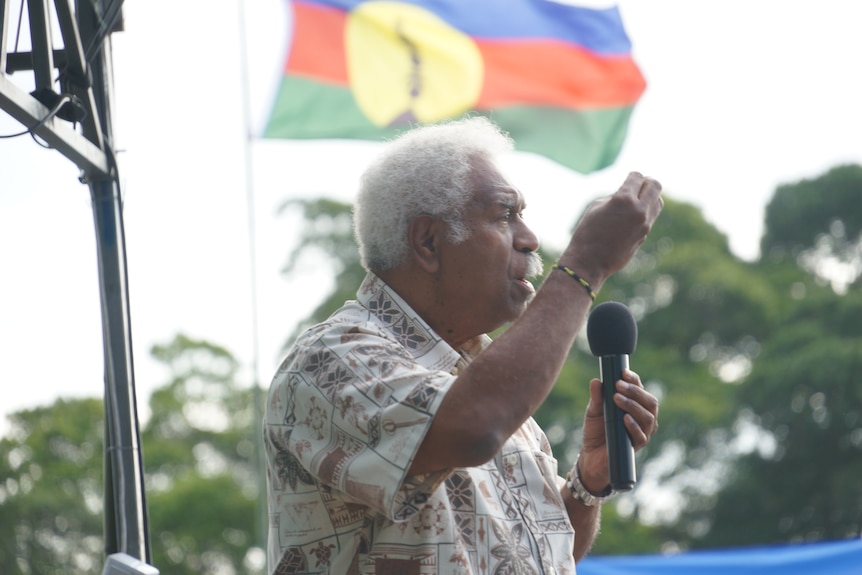 A man speaking to a crow with the Kanak flag in the background.