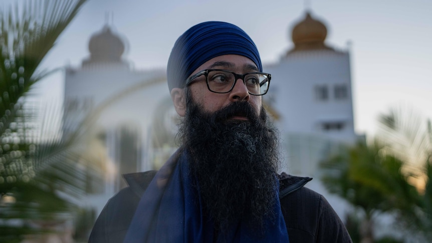 A man with a large beard wearing glasses and a turban looks to the side standing outside a Sikh temple.