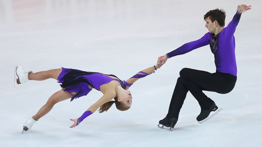 A male figure skater braces himself as he holds his female partner by one hand as she leans backward close to the ice. 